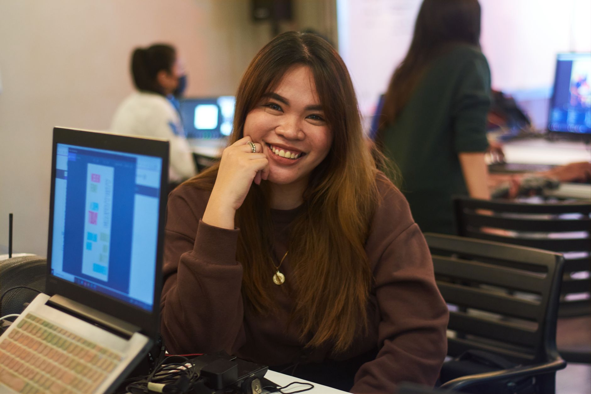 Asian woman smiling while working at a desk with laptops in a busy office environment.
