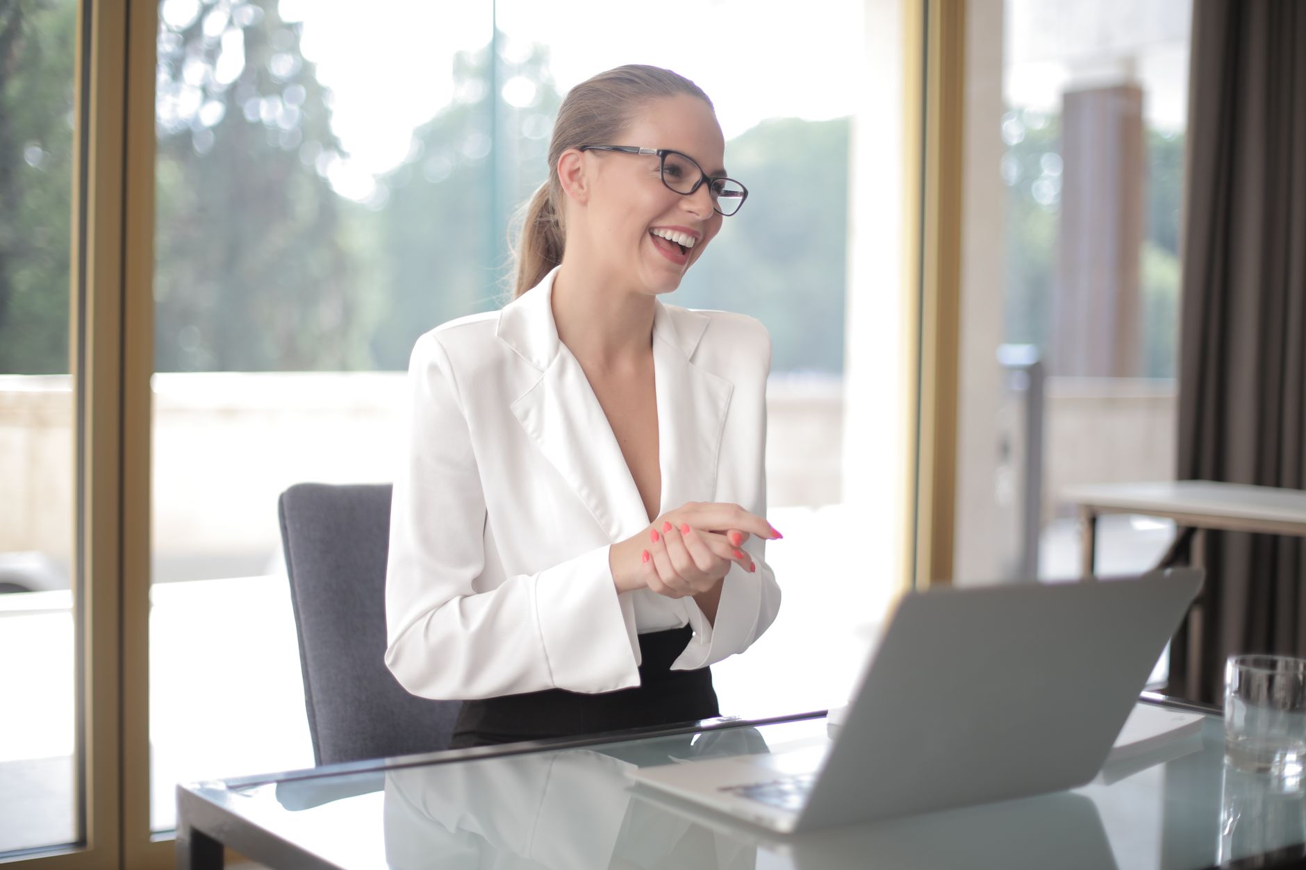 Woman entrepreneur in formal wear and eyeglasses sitting at table with laptop and documents and looking away while working on project in modern office